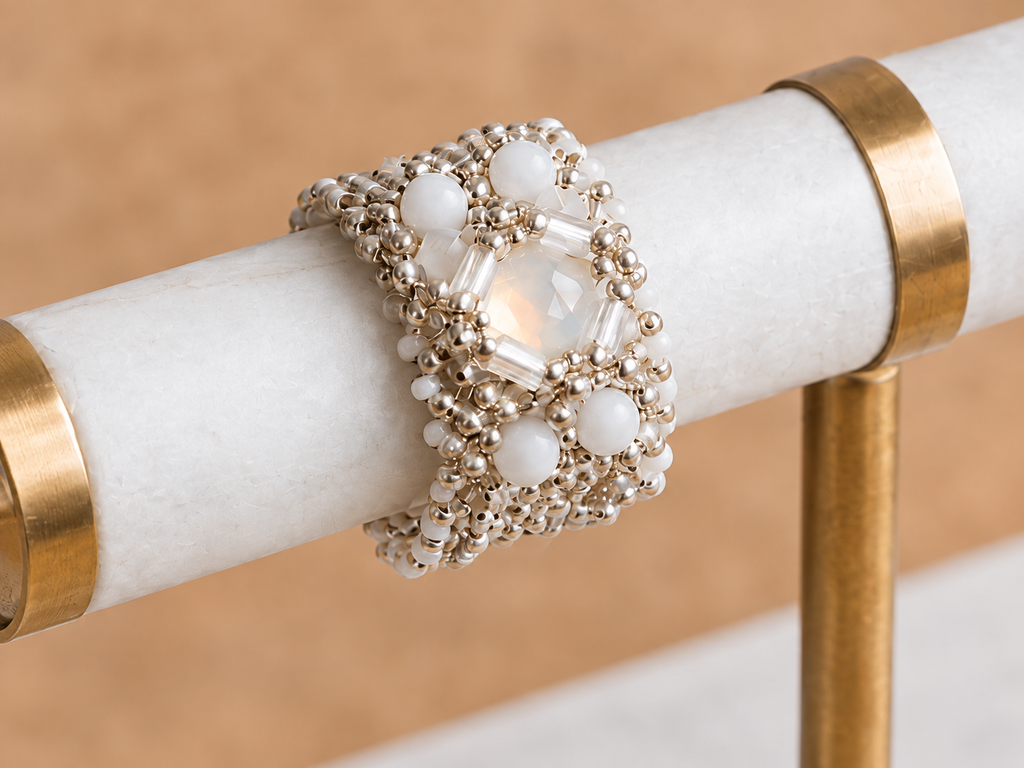 A wide beaded cuff ring with white faux pearls and clear faceted crystals, displayed on a white marble ring holder with gold bands.